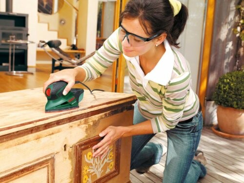 Woman using an electric sander on wooden furniture indoors.